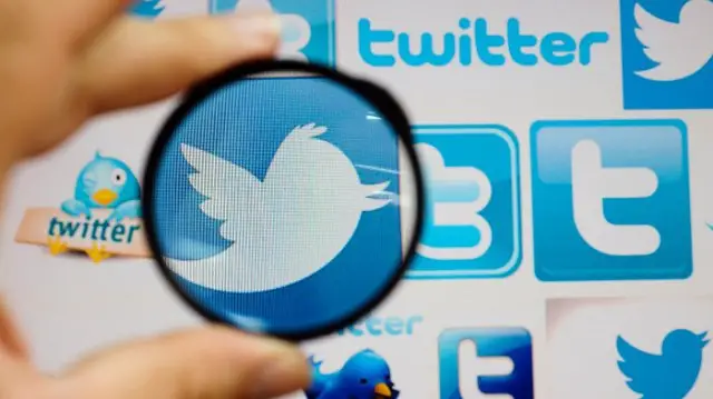 A person holds a magnifying glass over a computer screen displaying Twitter logos